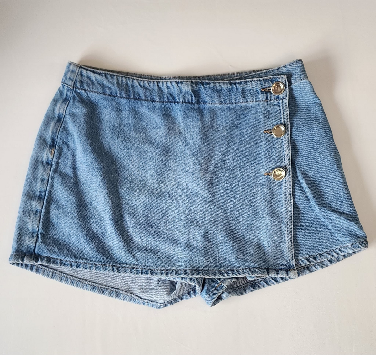 Style Reset light wash denim skort with a wrap-front design and metallic button detailing on a white background